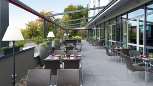 An outdoor patio with tables and chairs, large windows, and potted plants. - Leonardo Hotel Völklingen - Saarbrücken (Voelklingen)