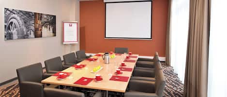 Un salón de conferencias con una larga mesa, sillas, una pantalla de proyector y un cuadro en la pared.