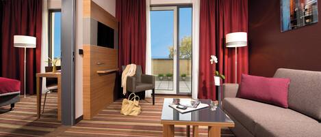 Une chambre d’hôtel équipée d’un canapé, d’une table basse, d’un fauteuil, d’une télévision, d’un bureau et d’une grande fenêtre avec des rideaux.