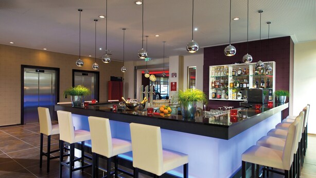 A modern bar area with a central island, bar stools, and hanging pendant lights. - Leonardo Hotel Völklingen - Saarbrücken (Voelklingen)