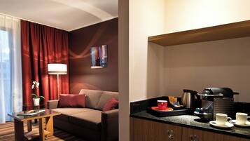 A hotel room with a sofa, a glass table, a lamp, a painting on the wall, and a kitchenette with coffee-making equipment.