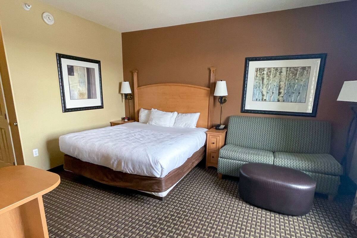 A hotel room with a large bed, two bedside tables with lamps, a green sofa, and a brown ottoman.
