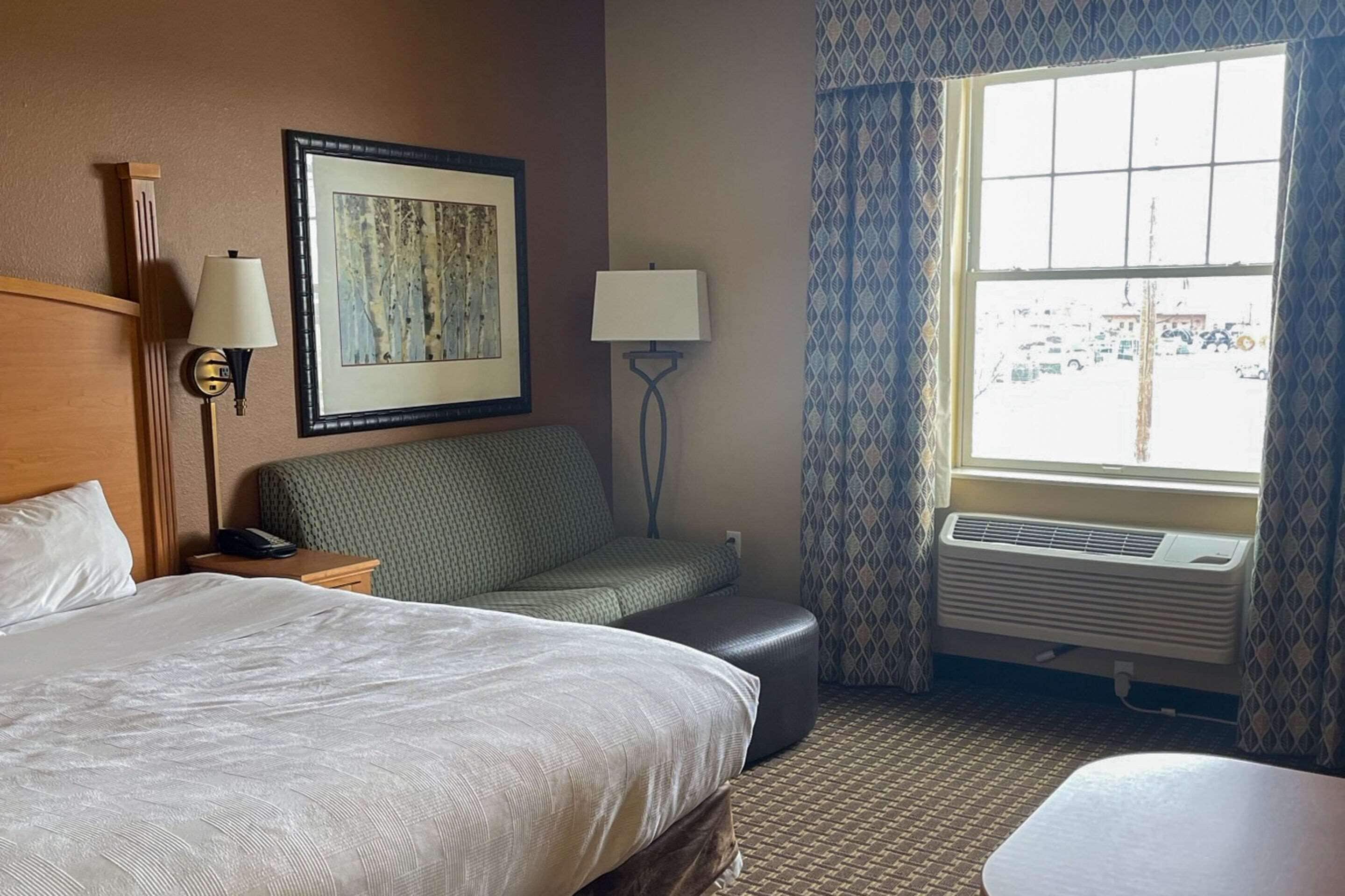 A hotel room with a large bed, a smaller sofa, a desk with a lamp, a framed picture on the wall, and a window with a view of a parking lot.