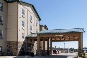 A hotel with a covered parking area, stone facade, and a sign indicating a clearance height of 13'6". - MainStay Suites Williston (Williston)