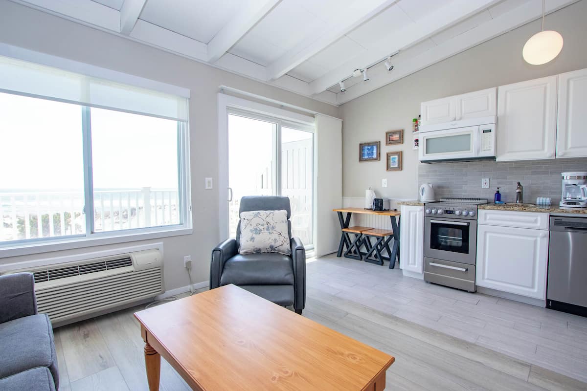 A modern kitchen with white cabinets, a built-in microwave, and an oven. There is a dining area with a table and chairs, and a window with blinds.