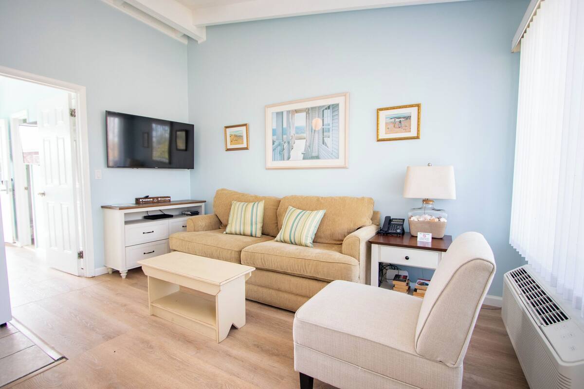 A living room with a beige sofa, a white armchair, a coffee table, a TV, and framed pictures on the wall.