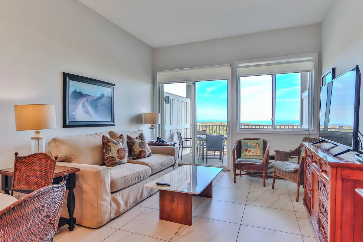 A living room with a sofa, coffee table, chairs, a TV, and a balcony with a view.
