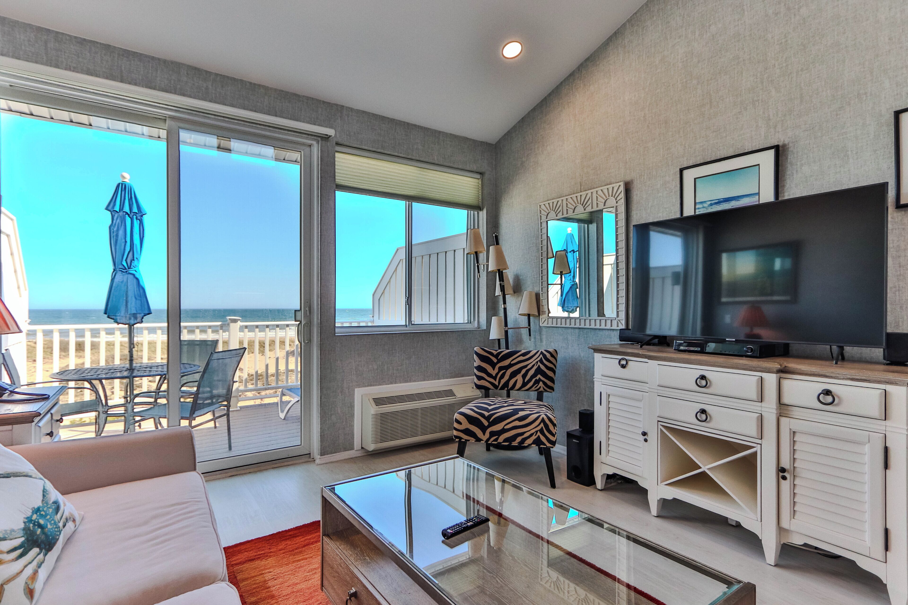 A living room with a glass coffee table, a zebra-patterned chair, a TV, and a view of the beach.