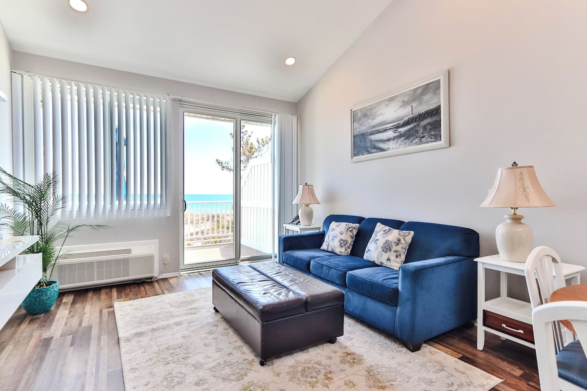 A living room with a blue sofa, a white lamp, a small table, and a view of the sea through large windows.