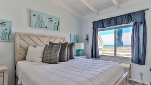 A bedroom with a bed, a nightstand, a lamp, artwork on the wall, and a window with curtains. - Sea Crest Resort (Amagansett)