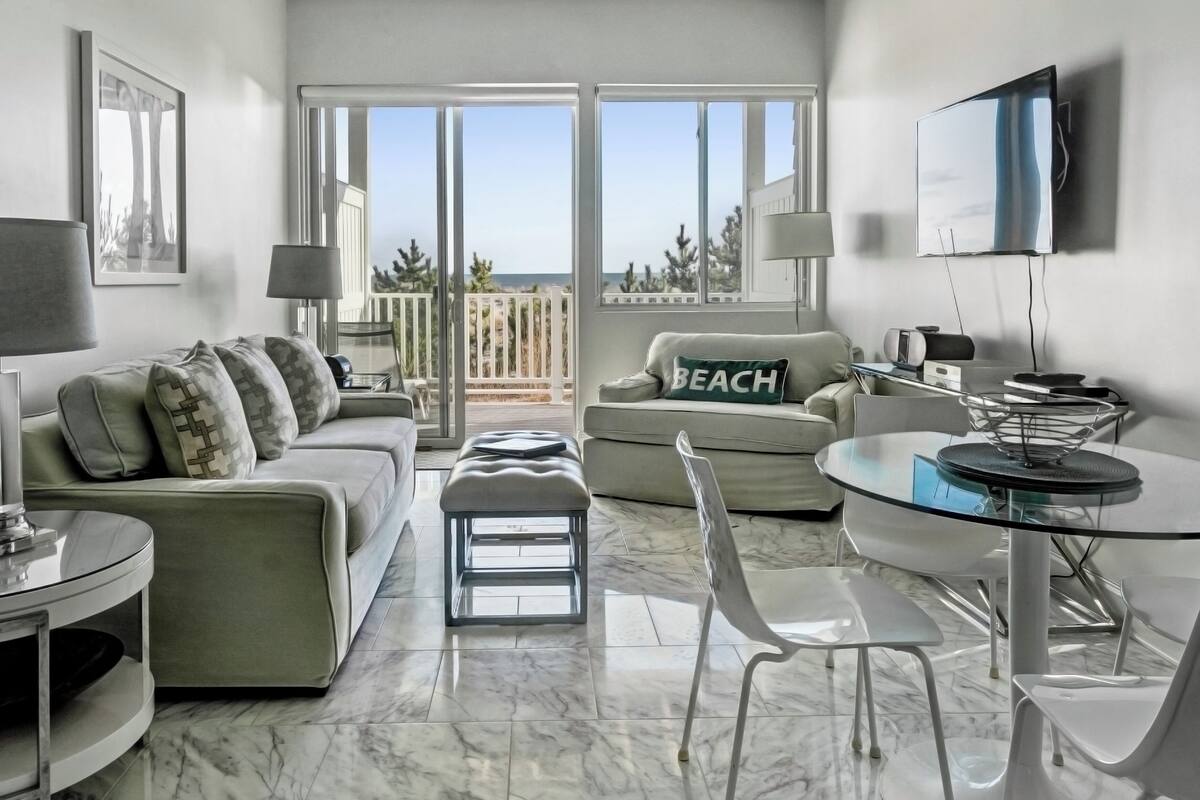 A modern living room with a sofa, ottoman, and a dining area with a glass table and chairs.