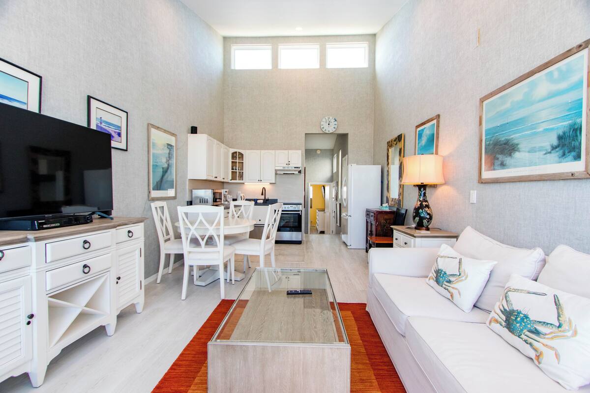 A modern living room with a glass coffee table, white furniture, and a kitchen in the background.