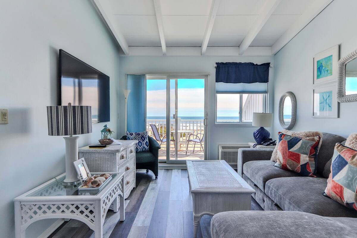 A room with a sofa, a table, a TV, and a view of the ocean through a sliding door.