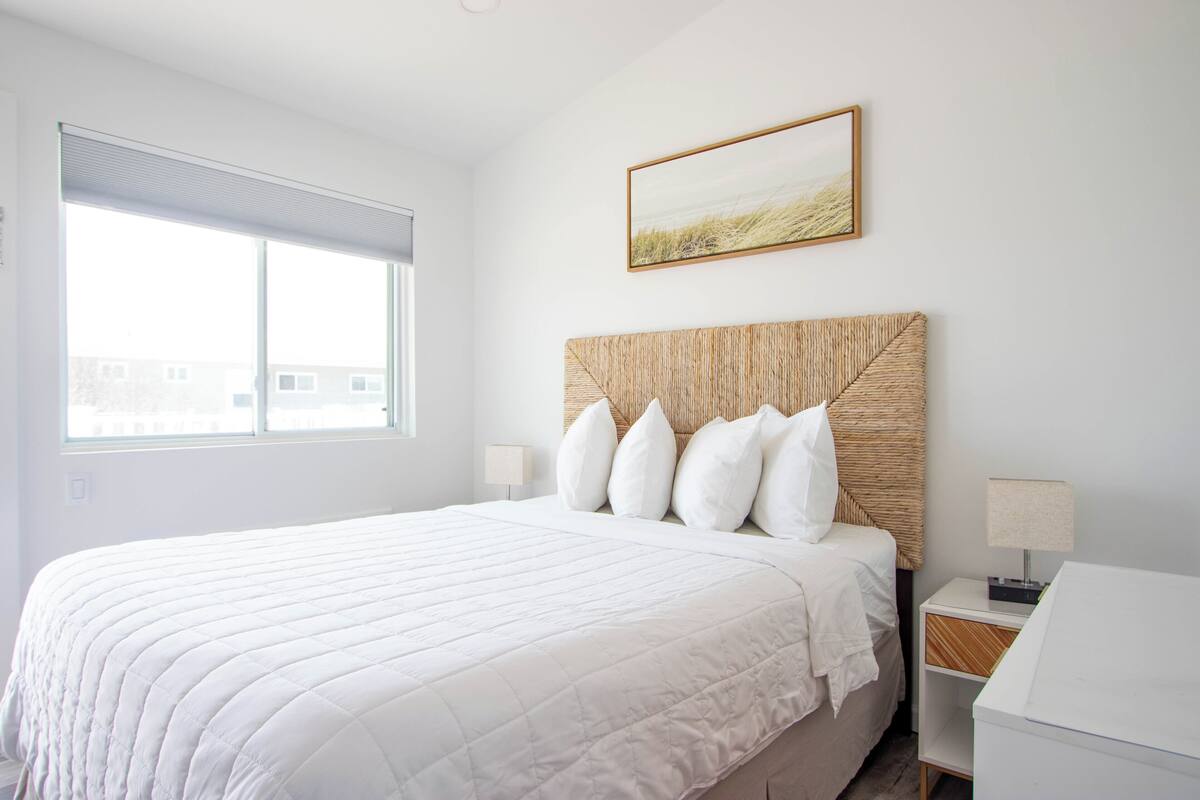 A bedroom with a large bed, a headboard, two bedside tables, a window, and a wall-mounted picture.