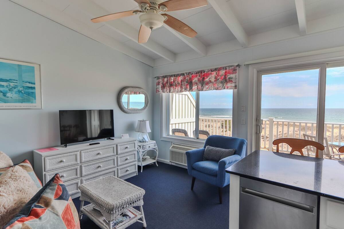 A room with a television, white dresser, blue armchair, and a view of the ocean.