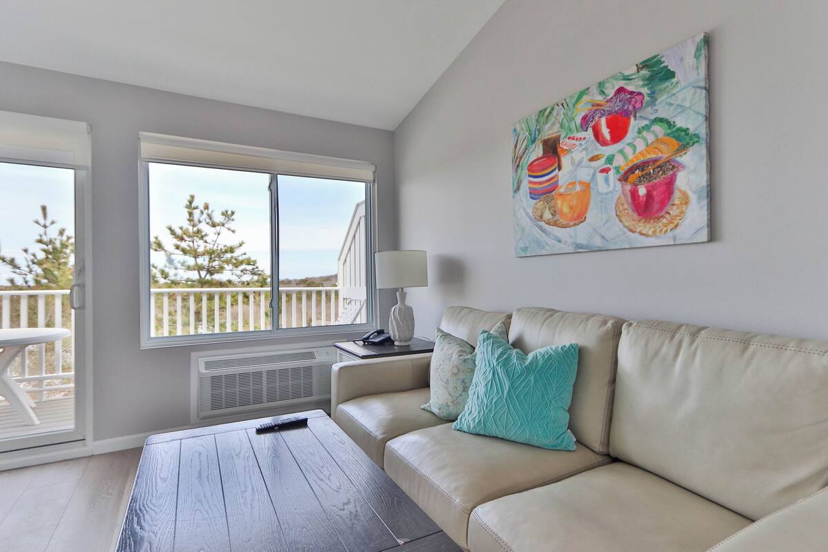 A living room with a beige sofa, a wooden coffee table, a painting on the wall, and a window with a view of trees and a balcony.