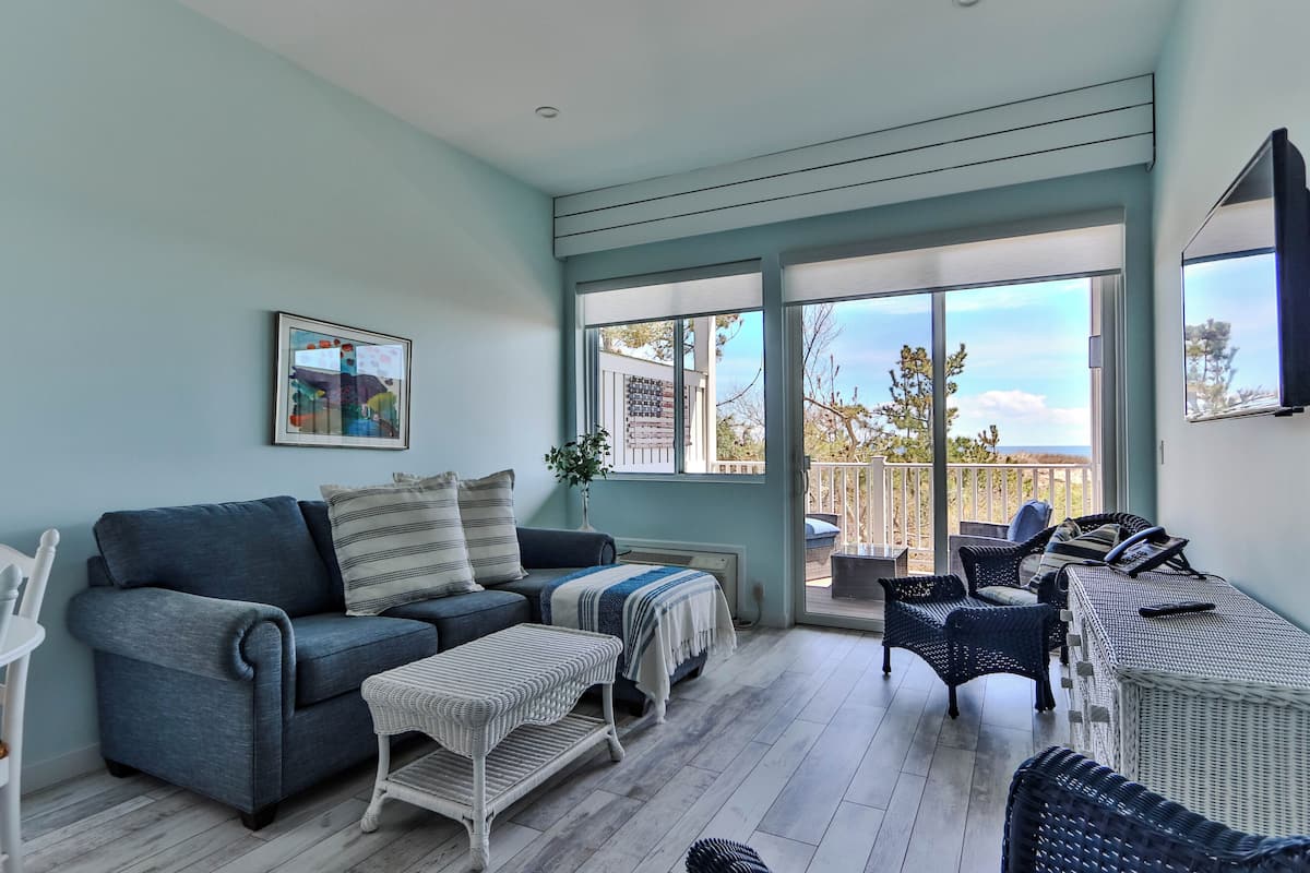A living room with a sofa, ottoman, wicker chairs, a table, and a large window with a view.