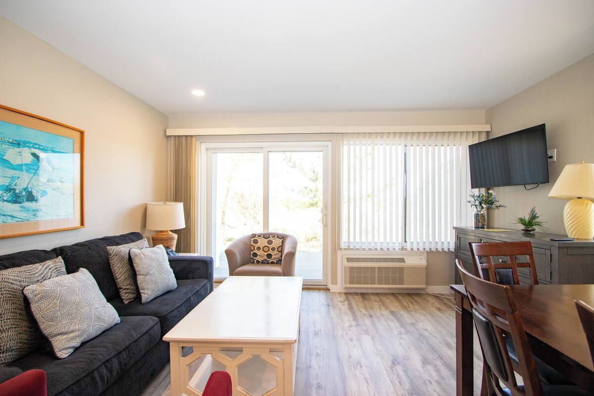 A living room with a sofa, a dining table, a TV, and a large window with blinds.