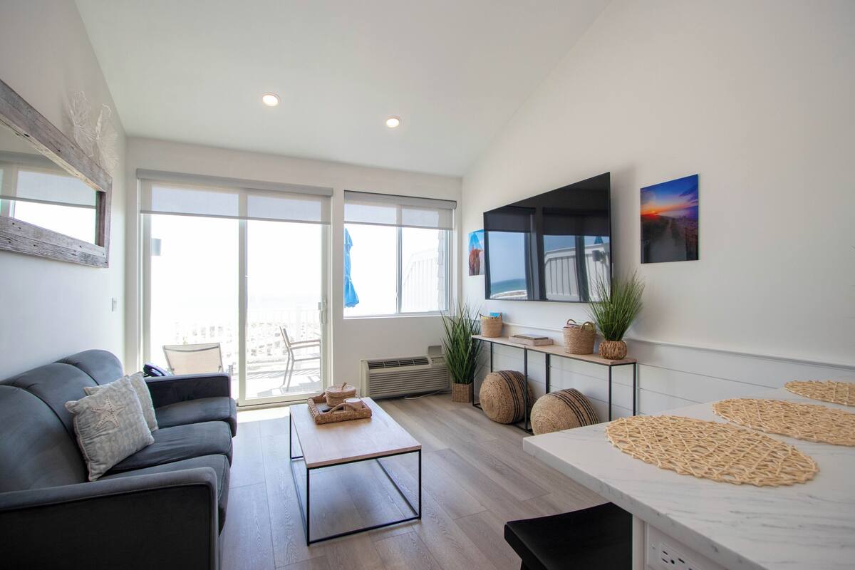 A modern living room with a sofa, a coffee table, a TV mounted on the wall, and large windows.