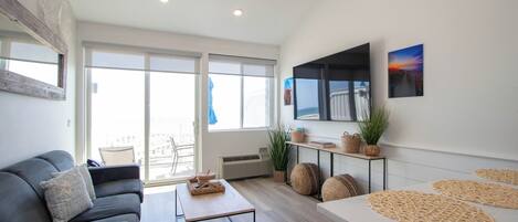 A modern living room with a sofa, a coffee table, a TV mounted on the wall, and large windows.