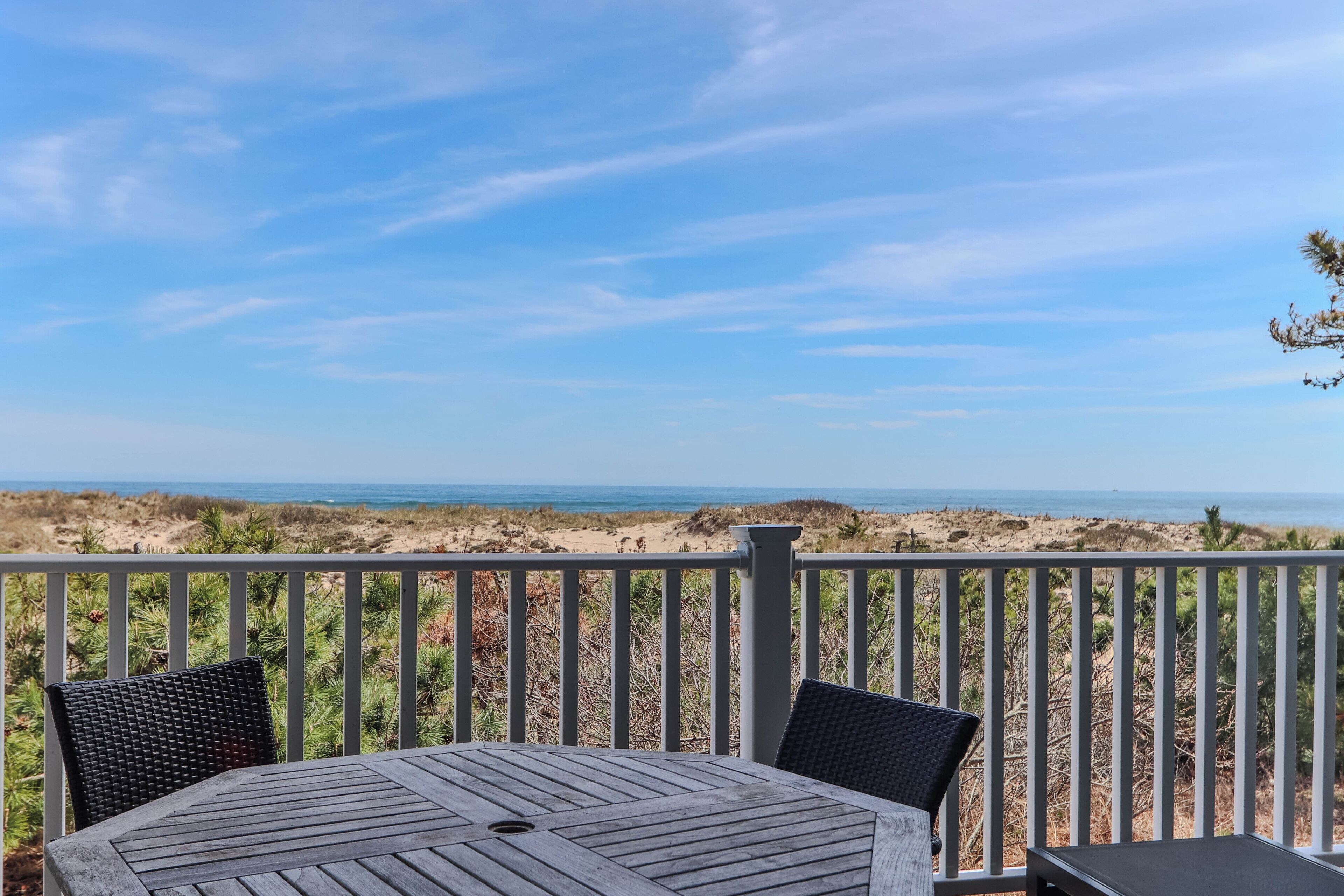 A wooden deck with a table and chairs, overlooking a beach with dunes and the ocean.