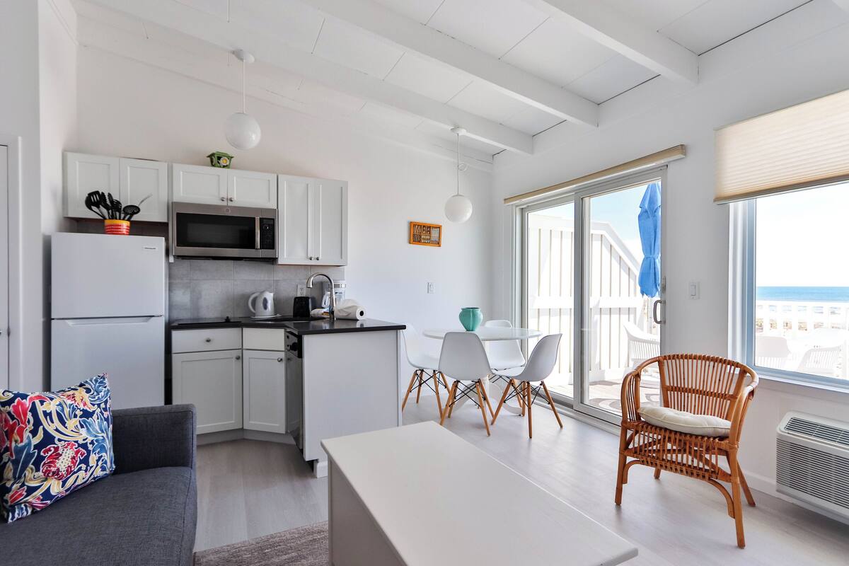 A modern kitchen with white cabinets, a refrigerator, and a microwave. There is a dining area with a table and chairs, and a wicker chair near a large window.