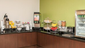 Una barra de desayuno con una máquina de café, un dispensador de zumo y diversas opciones de comida como frutas y cereales.