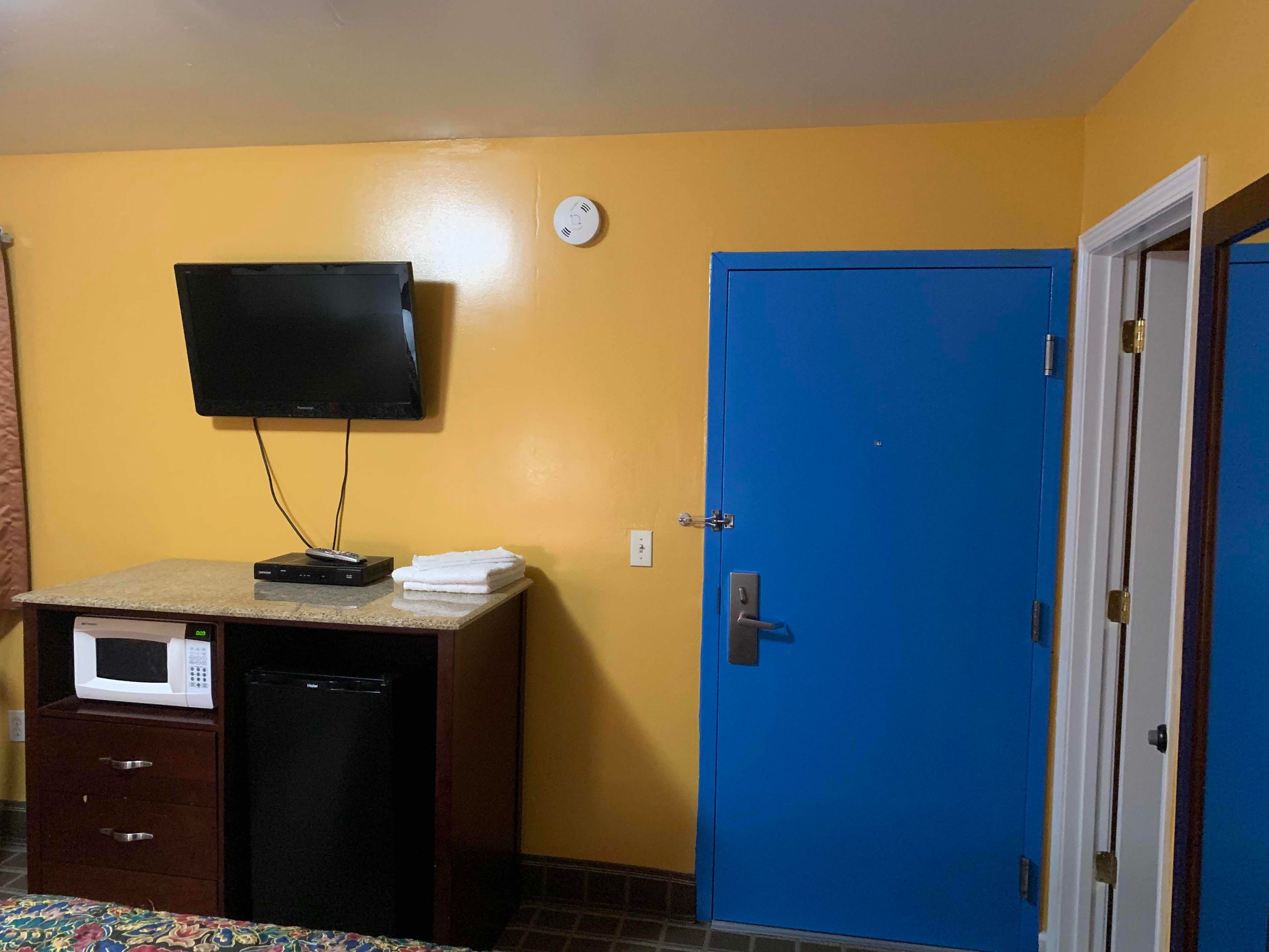 A room with a television mounted on the wall, a microwave on a counter, a blue door, and a small cabinet with drawers.