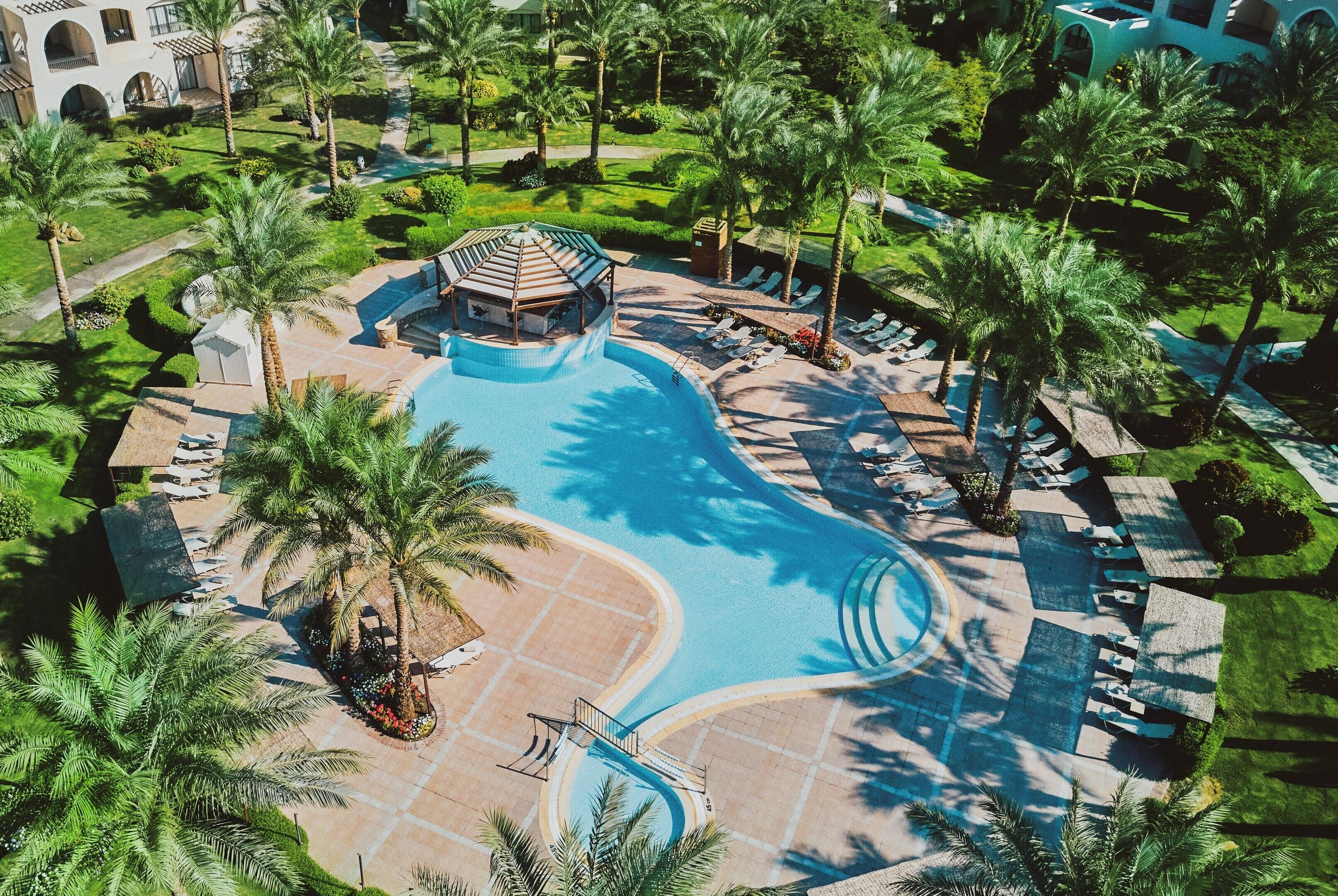 An aerial view of a resort with a central swimming pool, palm trees, and lounge areas.