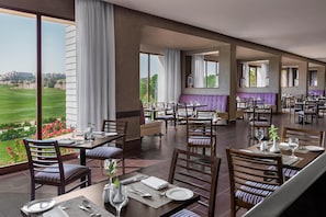 A restaurant with multiple dining tables and chairs, large windows with curtains, and a view of a green landscape.