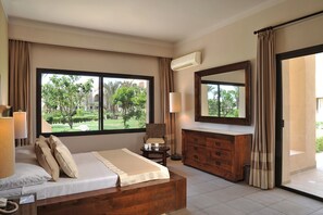 A hotel room with a large bed, a wooden dresser, a chair, and a view of a garden through a large window.