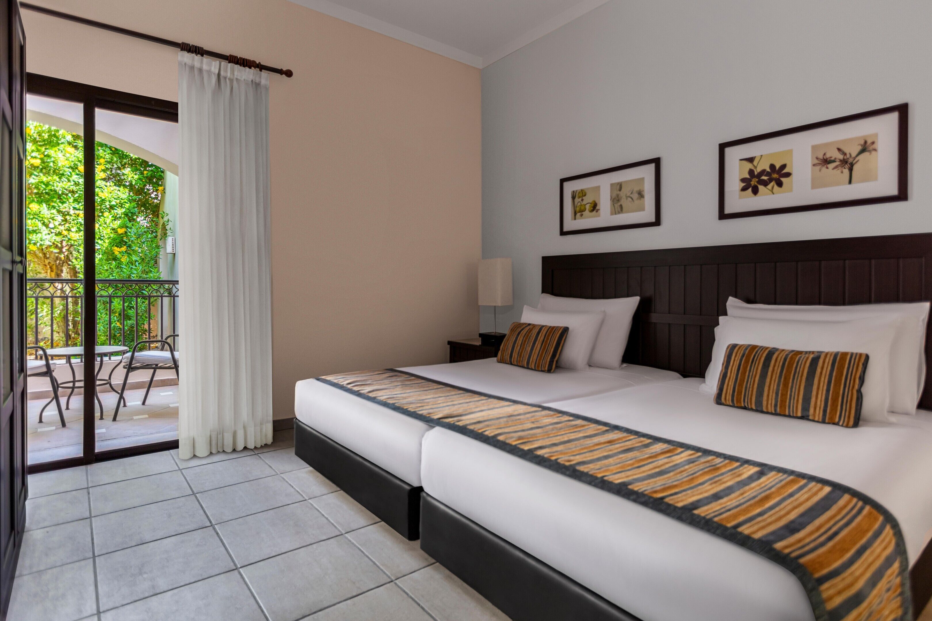 A hotel room with two beds, a balcony, and a view of greenery.