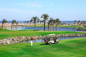A golf course with a water hazard, palm trees, and a golf cart. - JAZ Neo Saraya Palms - All inclusive (Makadi Bay)