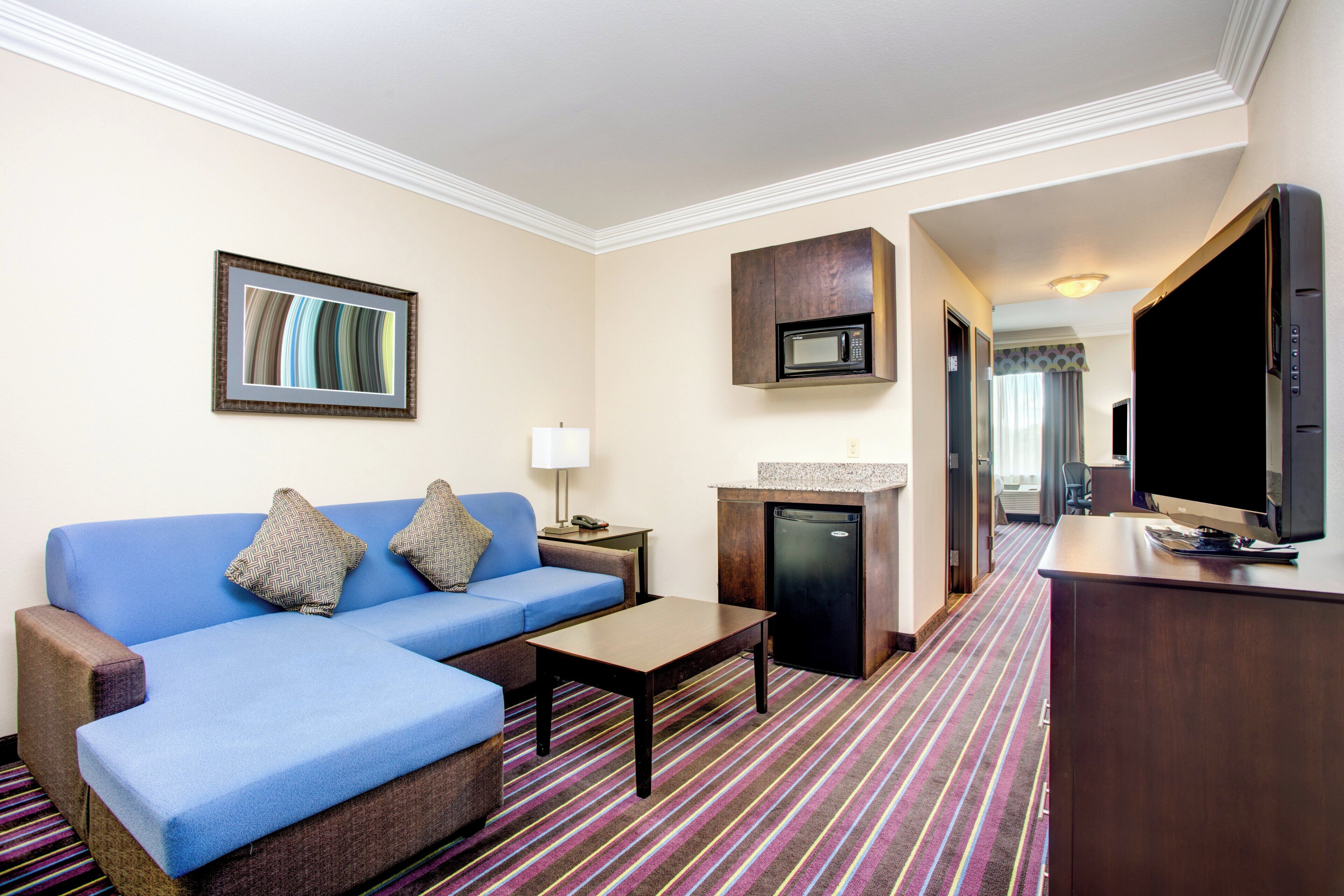 A hotel room with a blue sofa, a small table, a microwave, a refrigerator, a television, and a framed picture on the wall.
