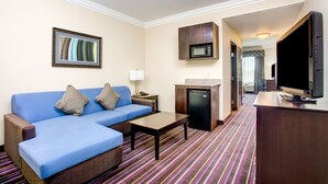 A hotel room with a blue sofa, a small table, a microwave, a television, and a kitchen area with a refrigerator.