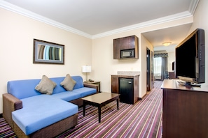 A hotel room with a blue sofa, a small table, a microwave, a television, and a kitchen area with a refrigerator. - Holiday Inn Express Hotel & Suites Raceland - Highway 90 by IHG (Raceland)