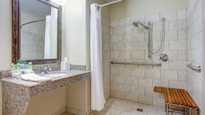 A bathroom with a granite countertop, a sink, a mirror, a shower with a grab bar, and a wooden bench.