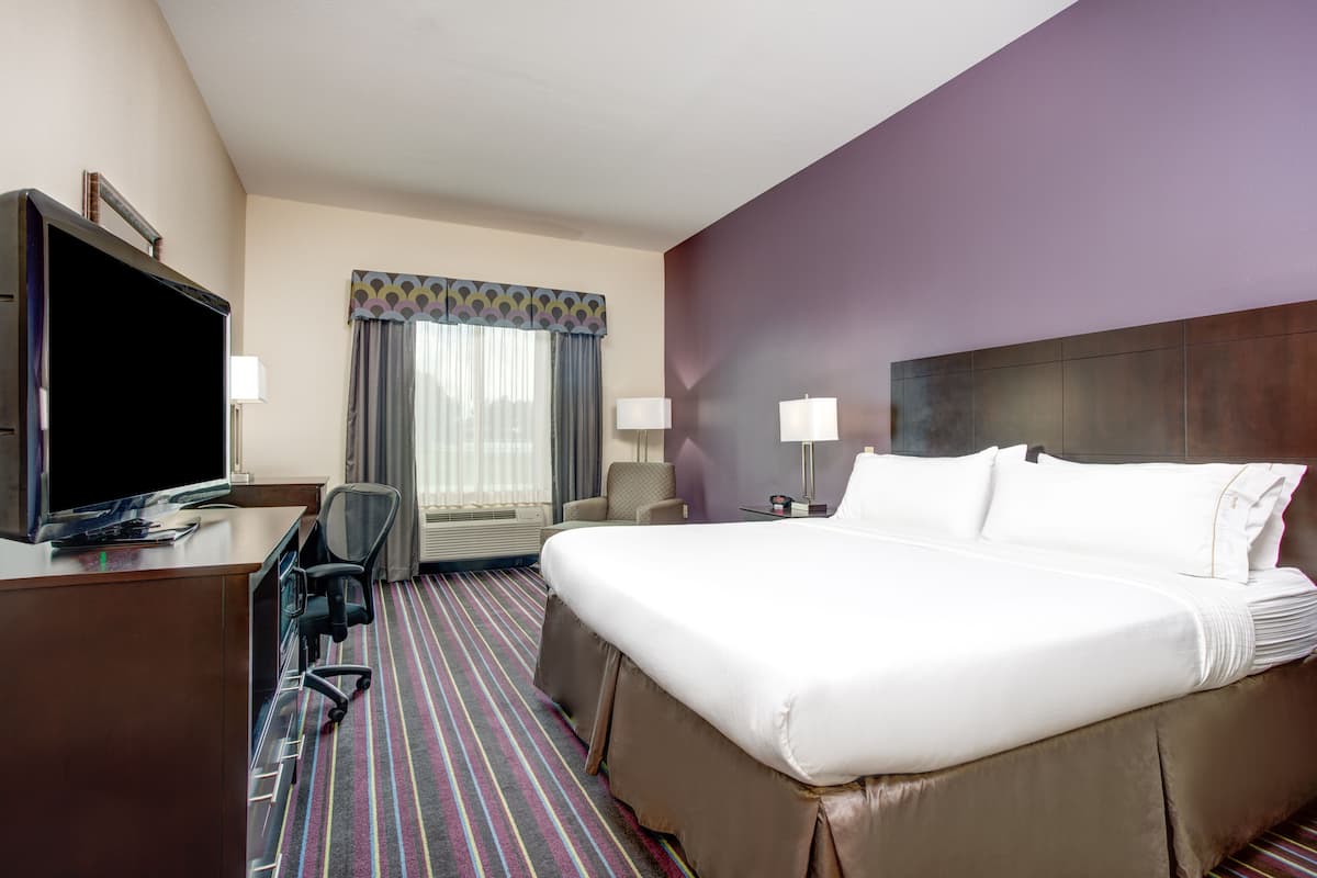 A hotel room with a large bed, a desk with a flat-screen TV, a chair, a lamp, and a window with curtains.