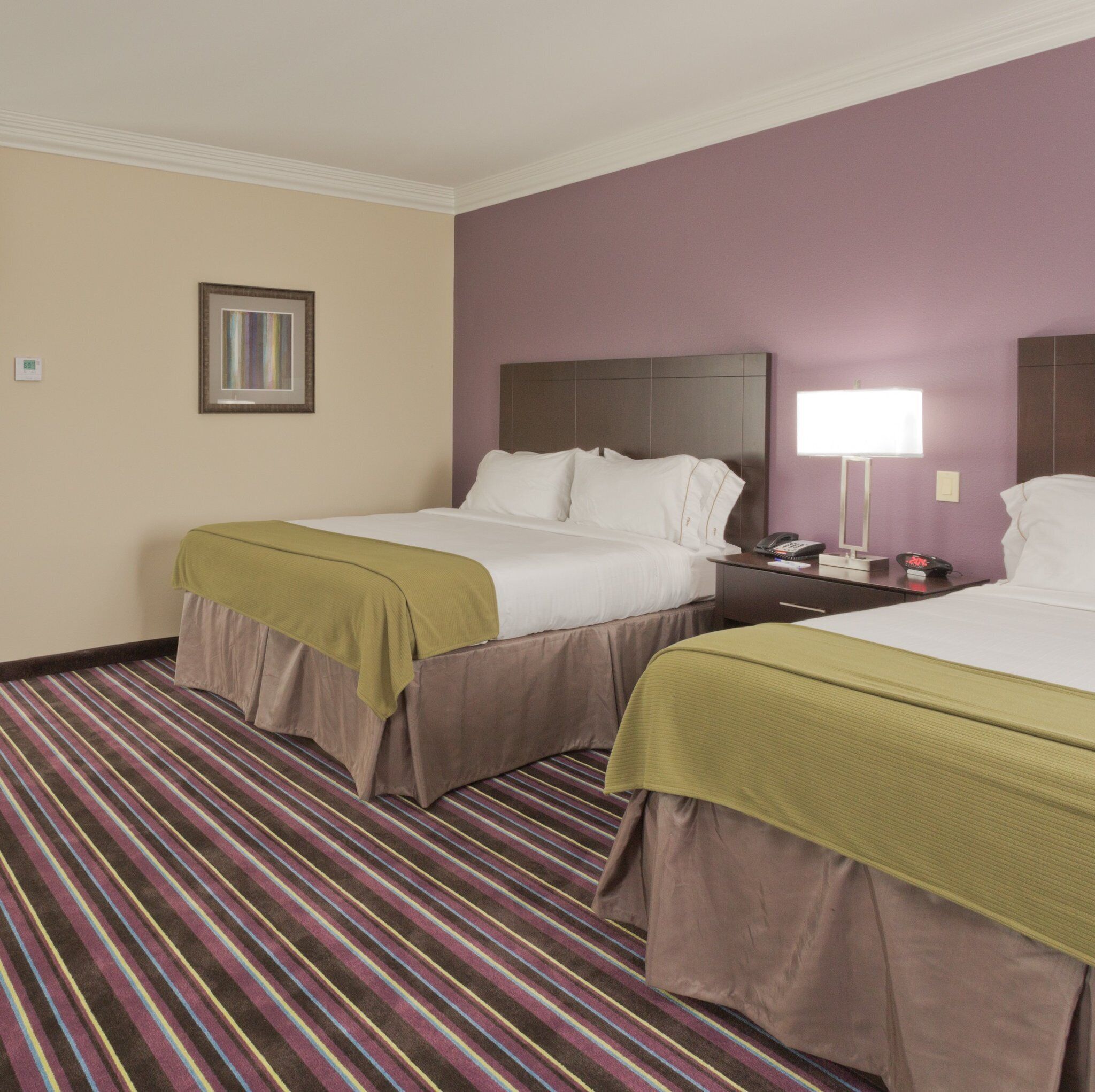 A hotel room with two beds, a striped carpet, a nightstand with a lamp, and a framed picture on the wall.