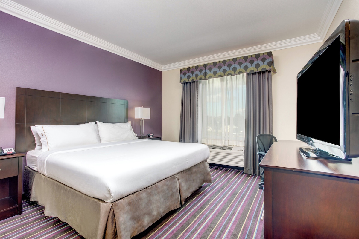A hotel room with a large bed, a desk with a flat-screen TV, a chair, and a window with curtains.