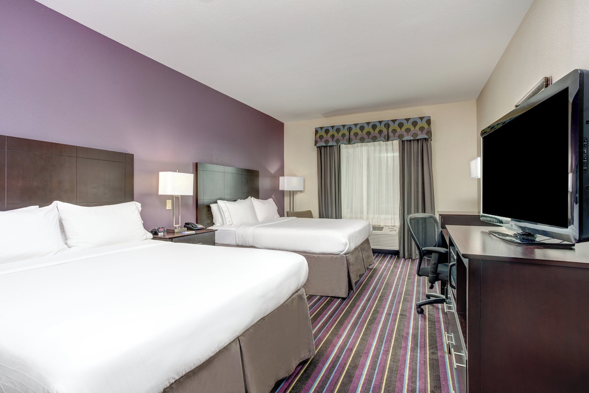A hotel room with two beds, a desk, a television, and a window with curtains.