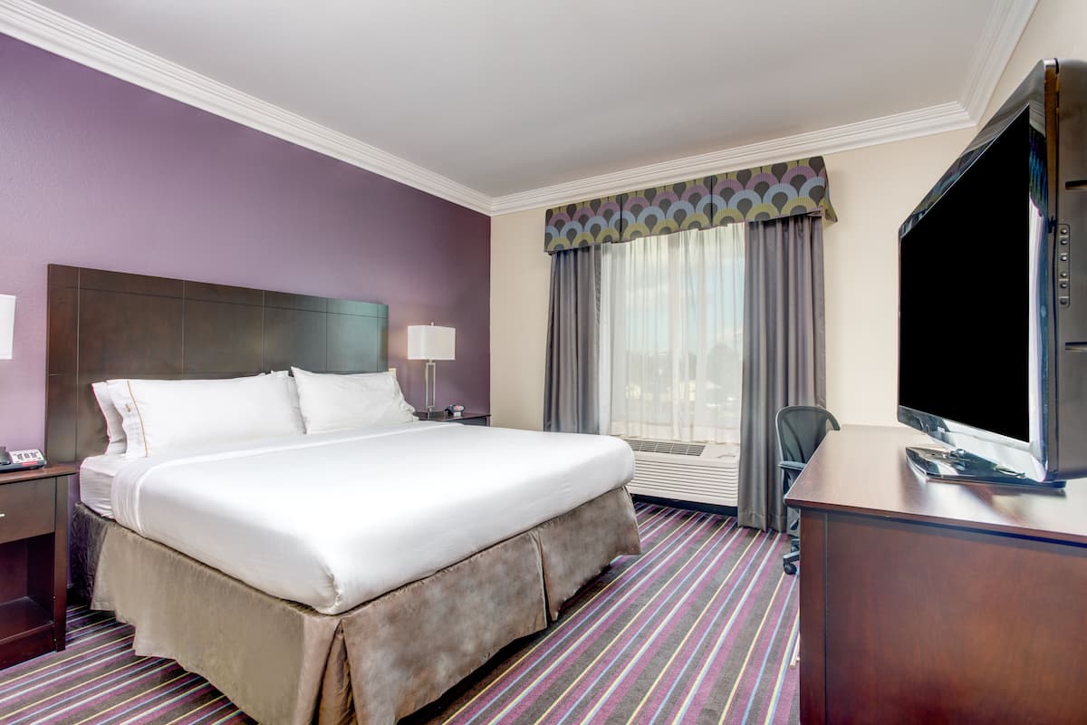 A hotel room with a large bed, a desk with a TV, a window with curtains, and a wall-mounted air conditioner.
