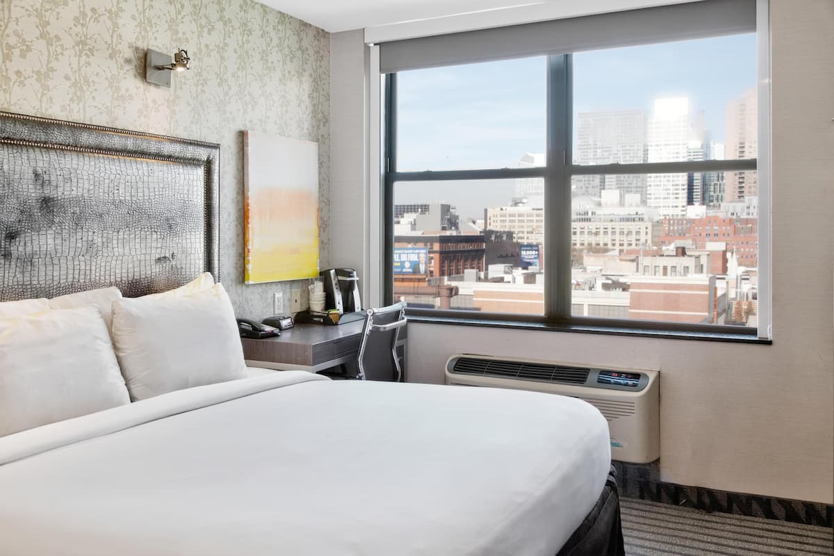 A hotel room with a large bed, a desk, a chair, a window with a city view, and a wall-mounted air conditioner.
