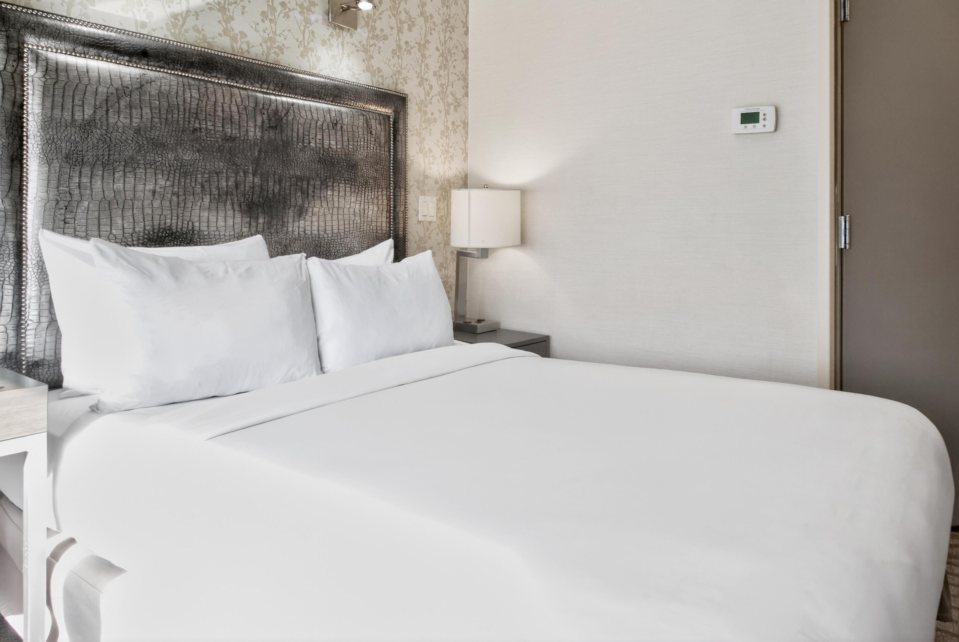 Ein Hotelzimmer mit einem großen Bett, einem Kopfteil mit Strukturmuster, einer Nachttischlampe und einem Thermostat an der Wand.