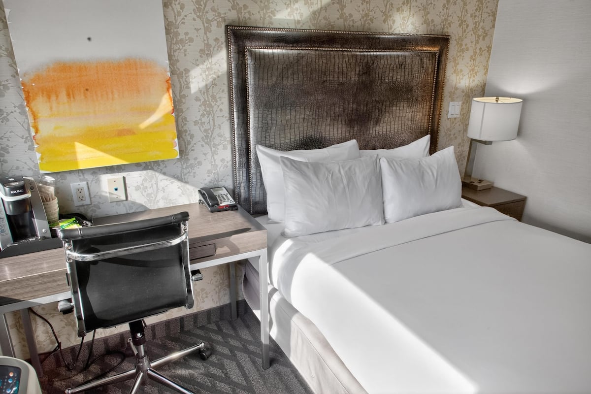A hotel room with a bed, a desk with a chair, a phone, a lamp, and a painting on the wall.