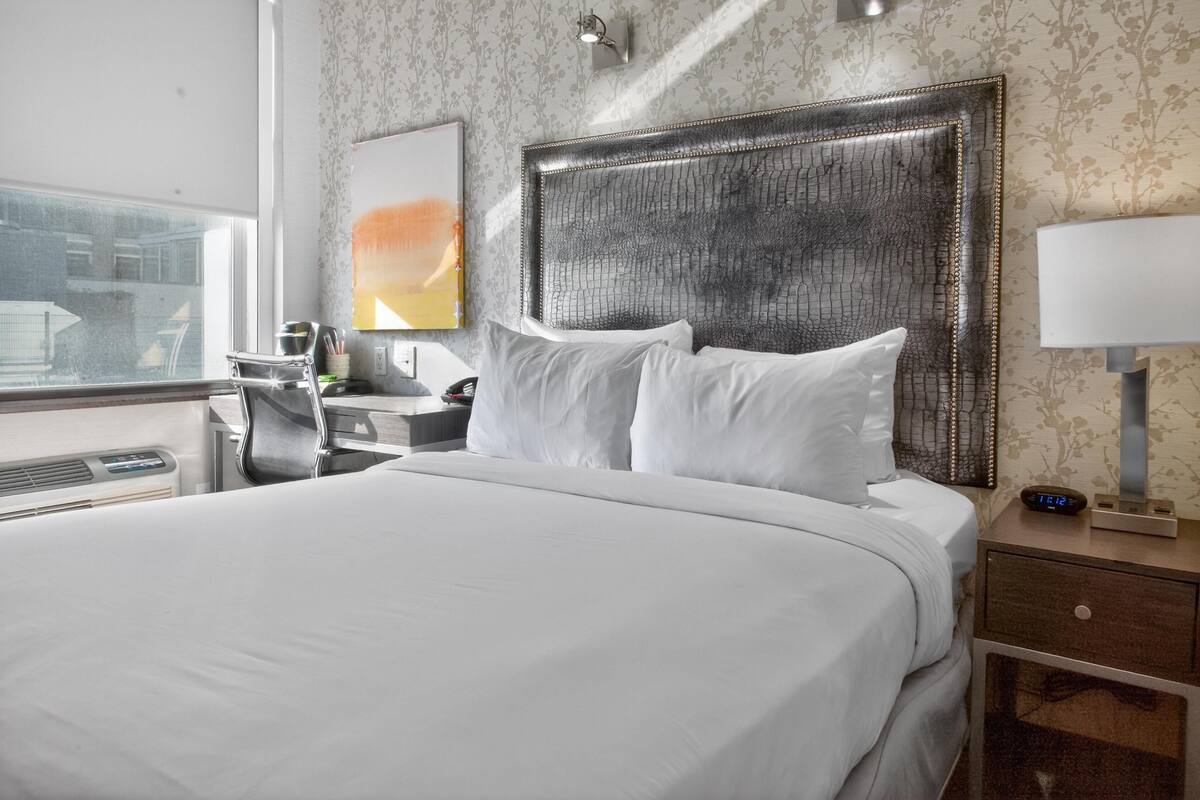 A hotel room with a large bed, a nightstand with a lamp, a clock, and a window with a view of buildings.