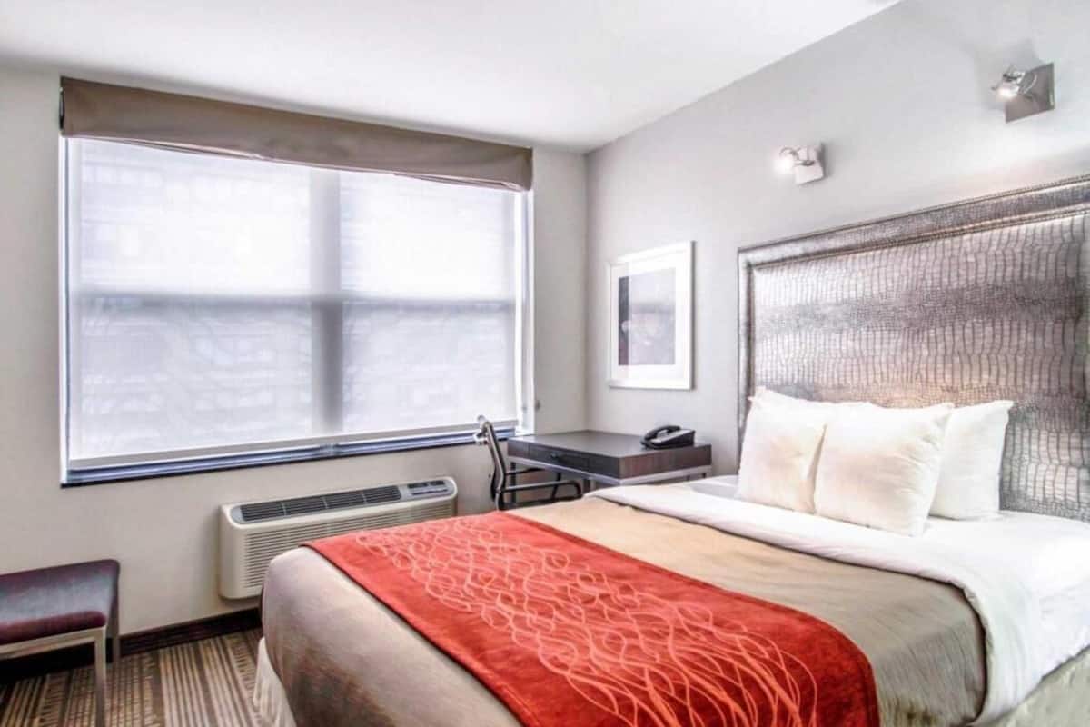 A hotel room with a large bed, a desk, a chair, a window, and a wall-mounted phone.
