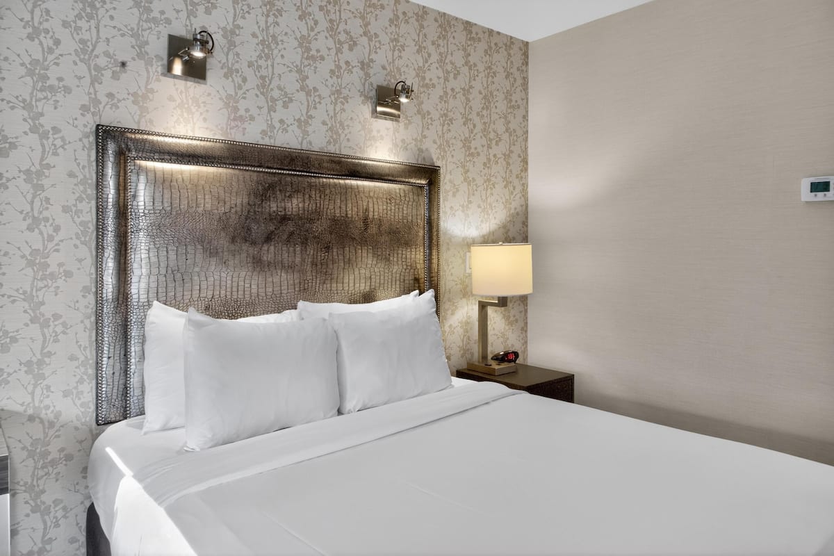 A hotel room with a large bed, a headboard with a textured design, a nightstand with a lamp, and a thermostat on the wall.