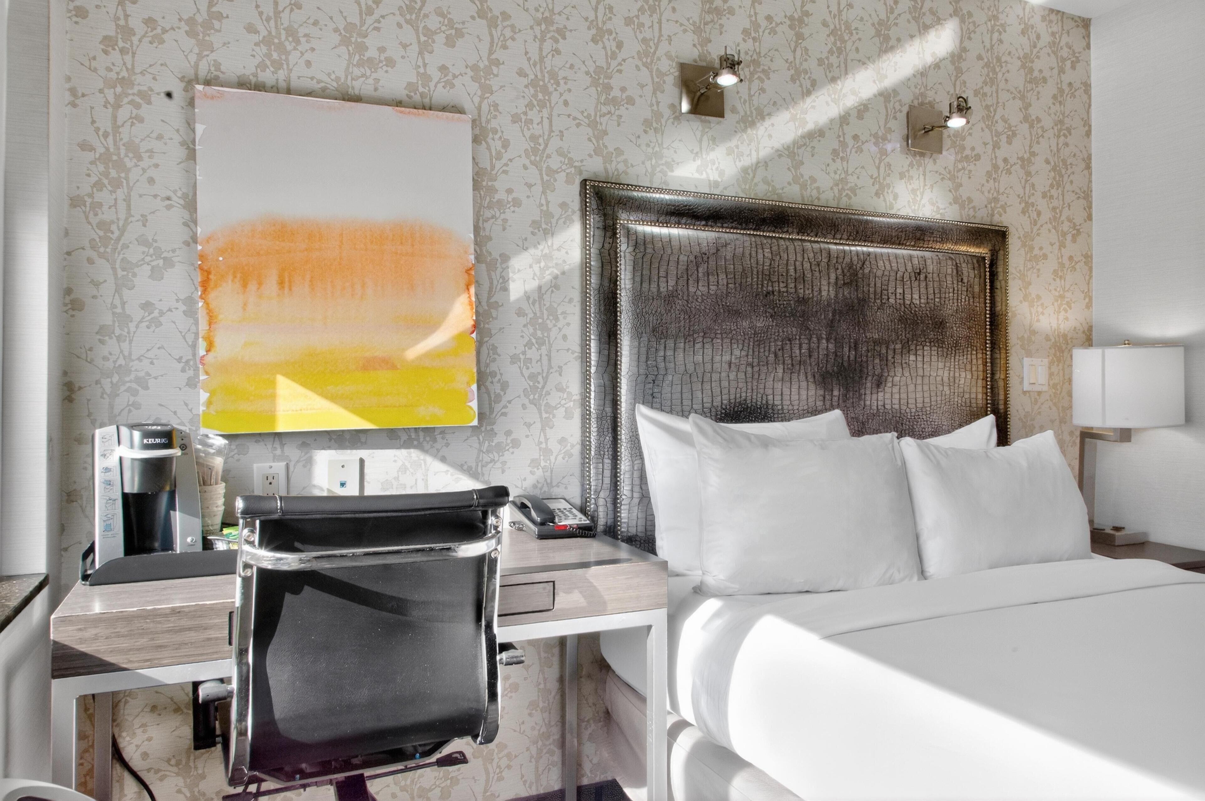 A hotel room with a bed, a desk with a coffee maker, a chair, and a painting on the wall.