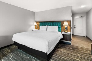 Una habitación de hotel con una cama grande, dos mesitas de noche con lámparas, una puerta y piso alfombrado.